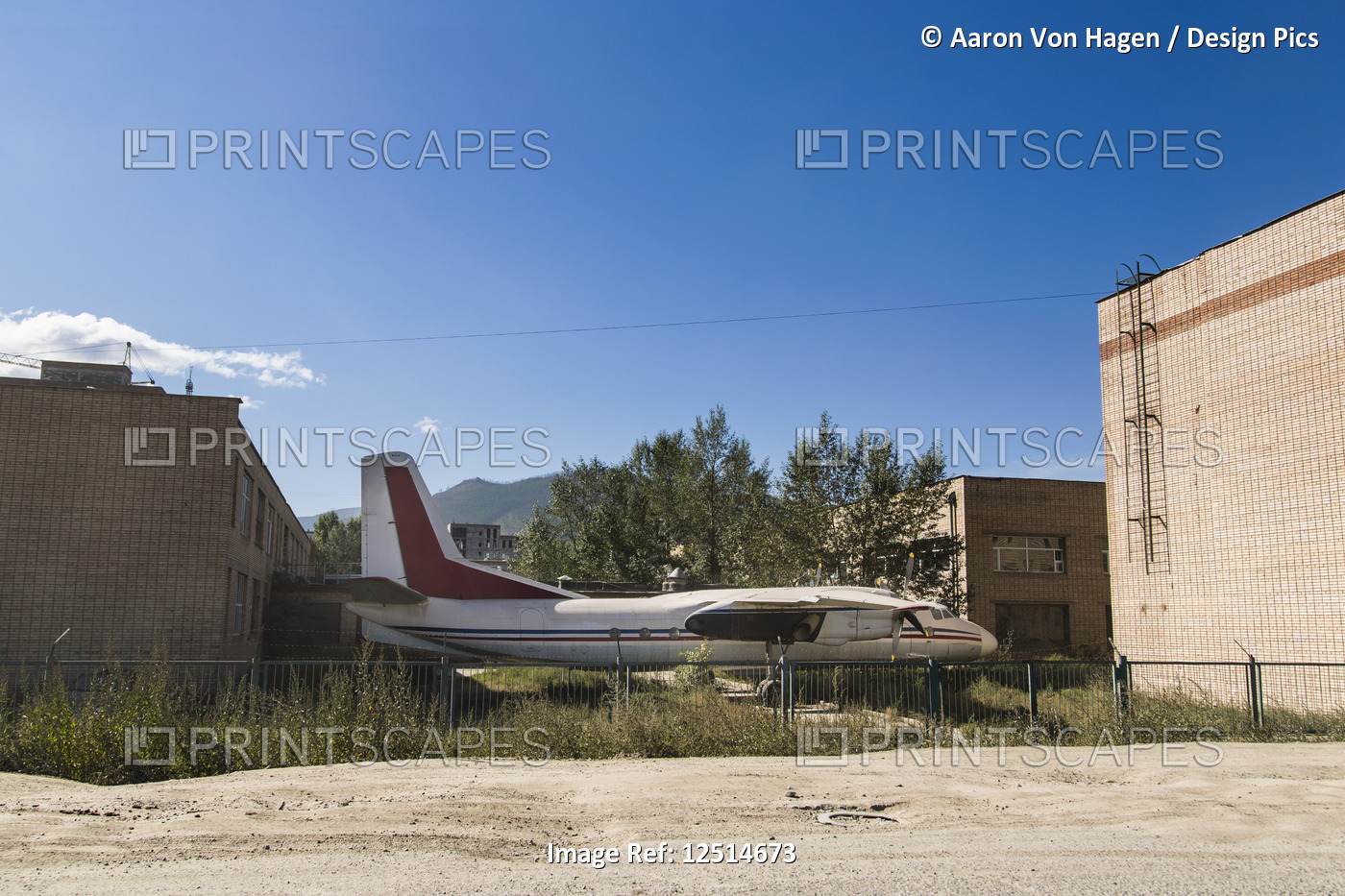 An old, abandoned plane parked between buildings and behind a fence; ...